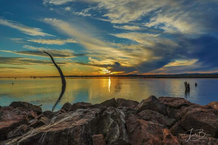 Tambo River Gippsland Australia as the sun is going down. It is popping out just a little before it finally sets. The clouds are whispy in the sky and there are dead trees in the water. The reflection on the water is amazing