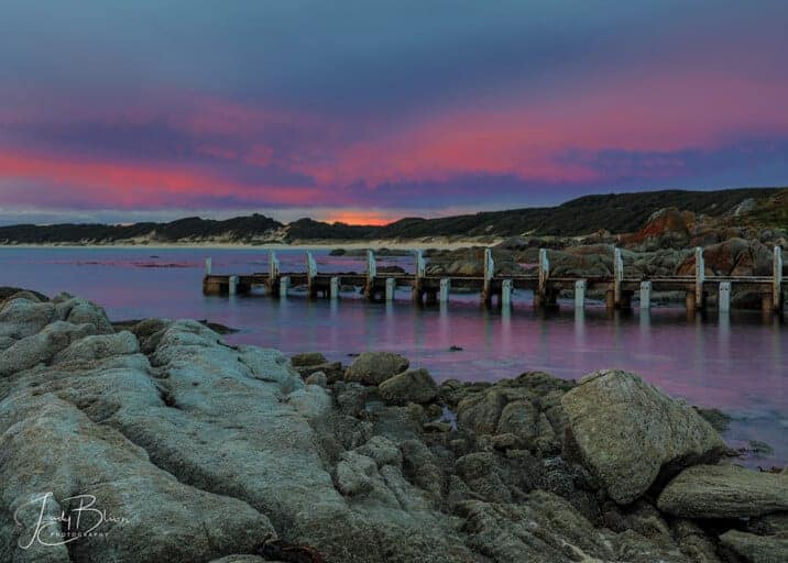 Cape Conron Gippsland Australia at sunset. The jetty is coming out of the water with the reflection of the purple and blue sky. The rocks are a stark grey against all of the colours in the picture